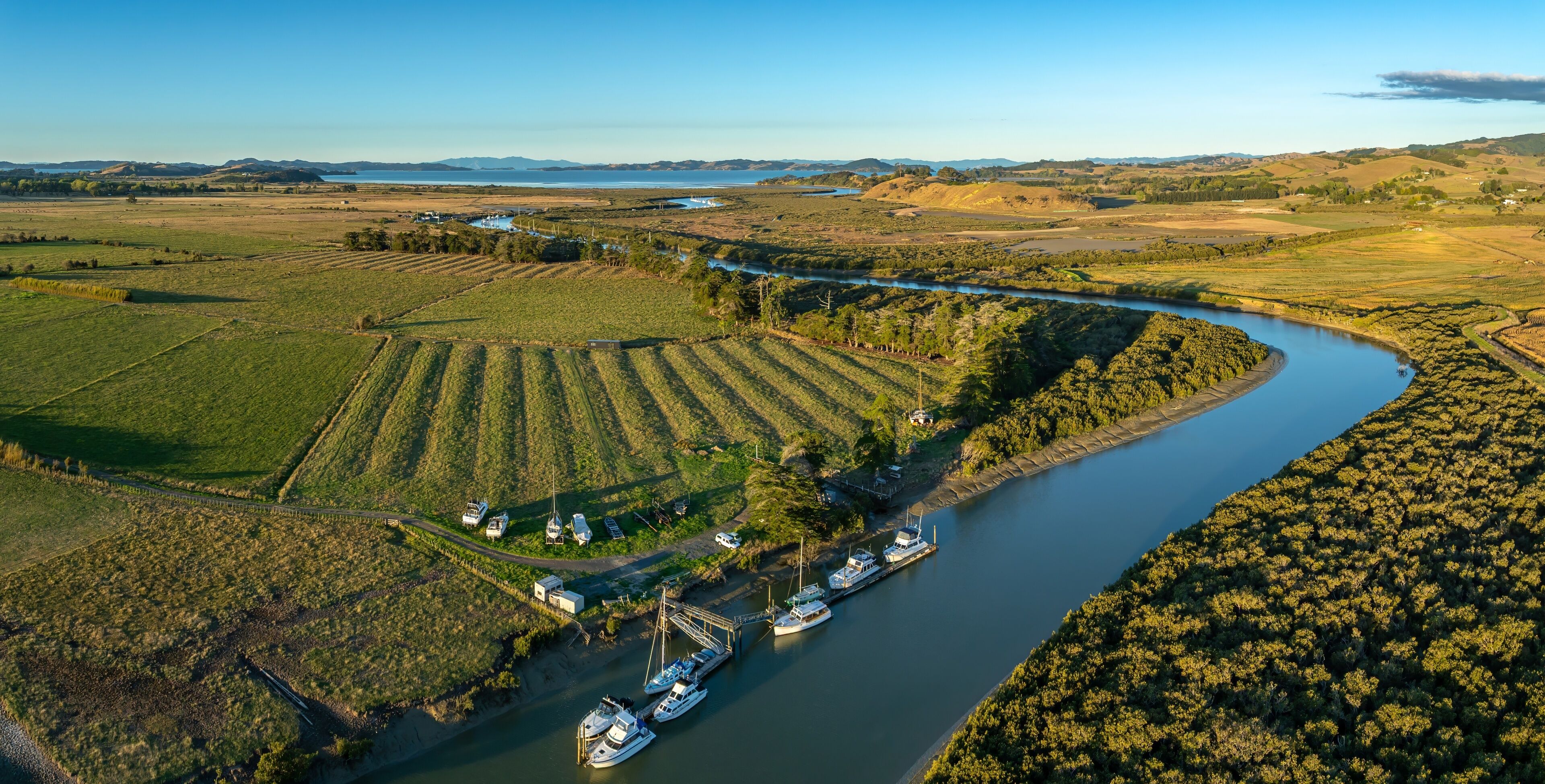Aerial view of boats docked on a river in a rural area of New Zealand. The boats are moored at a small pier, surrounded by farmland and trees. The river winds through the landscape. CLEVEDON, NZ