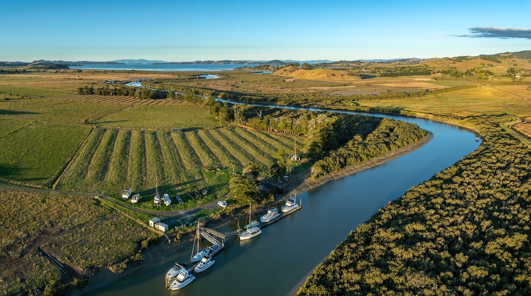 Aerial view of boats docked on a river in a rural area of New Zealand. The boats are moored at a small pier, surrounded by farmland and trees. The river winds through the landscape. CLEVEDON, NZ