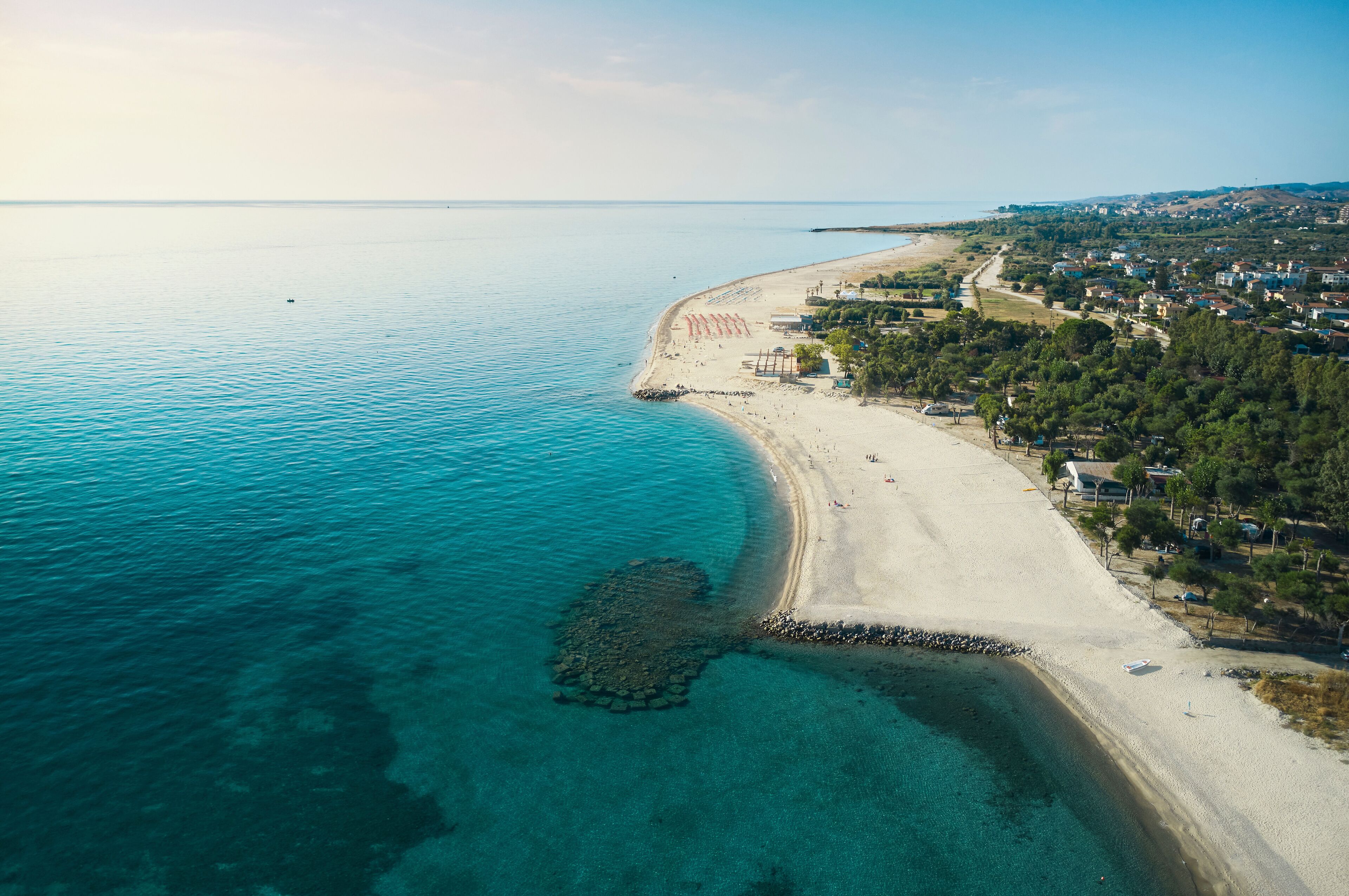 An aerial view of a coastal area with a sandy beach, clear turquoise waters, and a rocky outcrop near the shore.