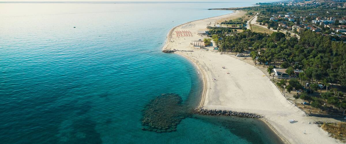 An aerial view of a coastal area with a sandy beach, clear turquoise waters, and a rocky outcrop near the shore.