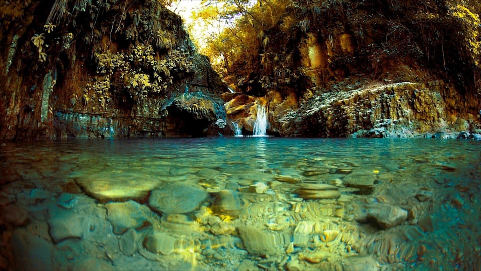 A cave with clear water, a waterfall, and rocky walls.