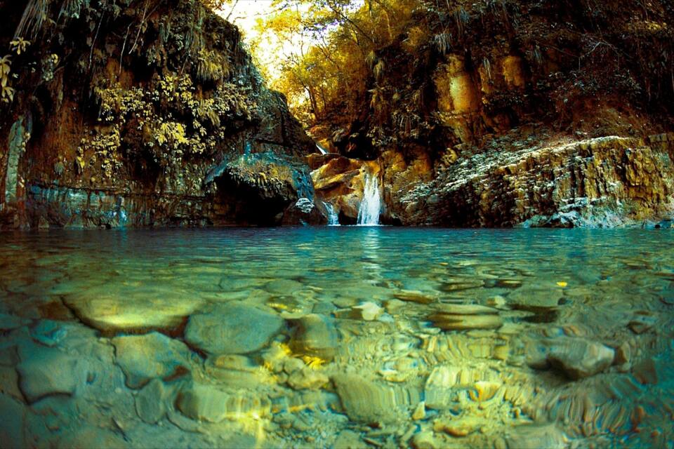 A cave with clear water, a waterfall, and rocky walls.