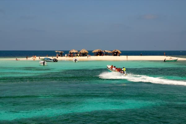 Ein tropischer Strand mit klarem türkisblauem Wasser, ein Boot, das über das Wasser gleitet, und strohgedeckte Hütten im Hintergrund.