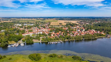 Een stad aan een meer, met huizen met rode daken, boten afgemeerd langs de oever en omliggende groene velden.