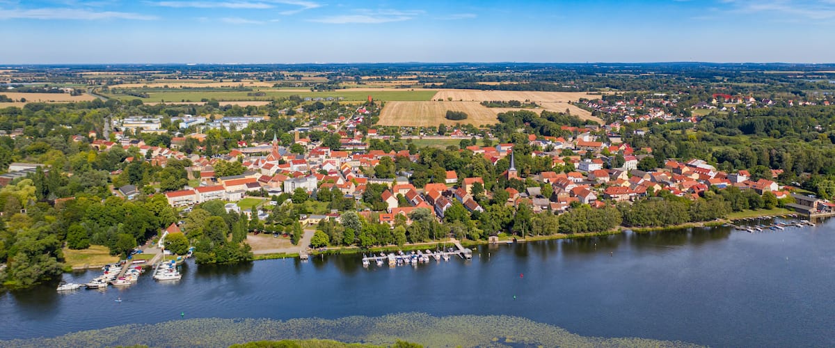 A lakeside town with red-roofed houses, boats docked along the shore, and surrounding green fields.