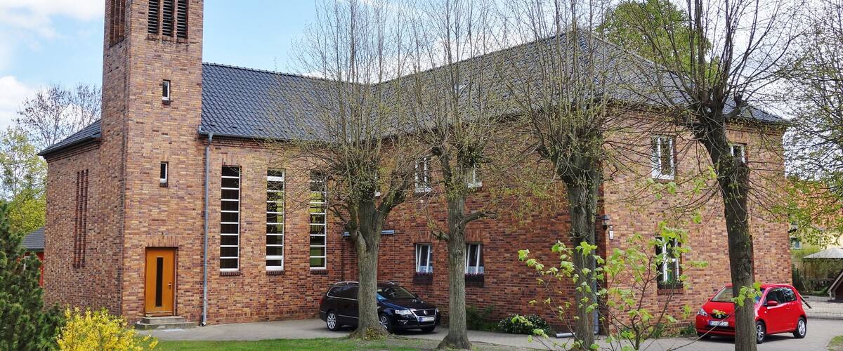 A brick church with a bell tower, two cars parked in front, and trees with green leaves.