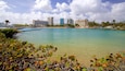 A coastal cityscape with clear turquoise waters, palm trees, and modern high-rise buildings.