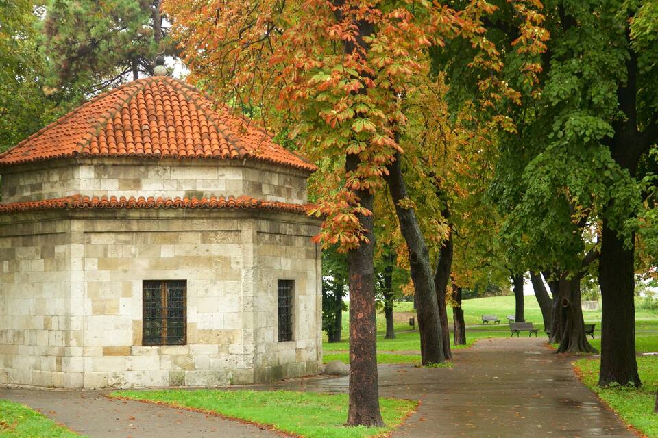 A small stone building with a red-tiled roof, surrounded by trees and a paved path.
