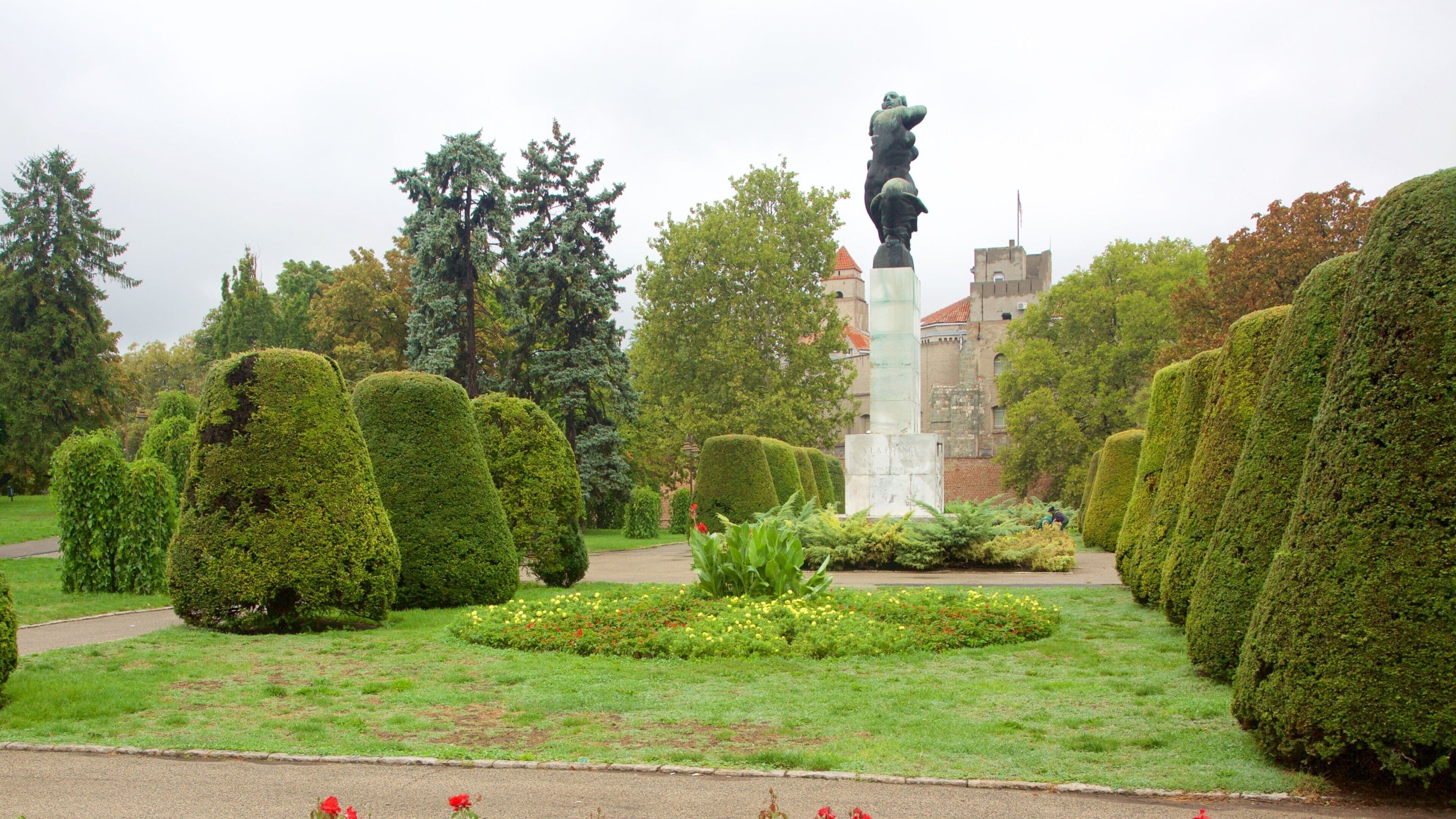 Una estatua en un parque con setos bien cuidados y un edificio al fondo.