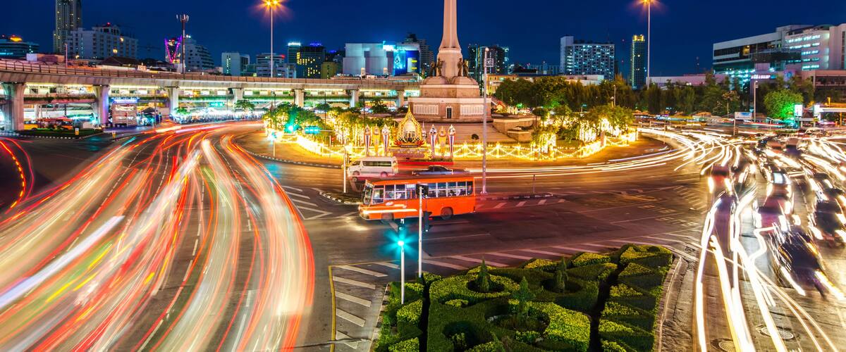 Un paisaje urbano nocturno con un monumento destacado, una intersección concurrida con estelas de luz de vehículos e edificios iluminados.