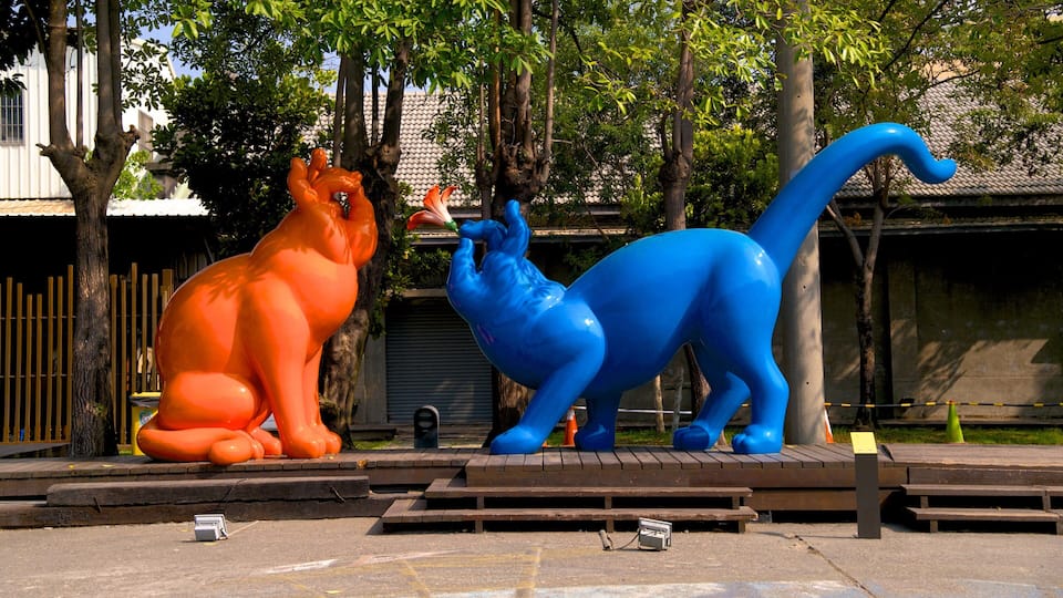 Duas grandes esculturas de animais coloridas, uma laranja e outra azul, sobre uma plataforma de madeira em ambiente externo, com árvores e edifícios ao fundo.
