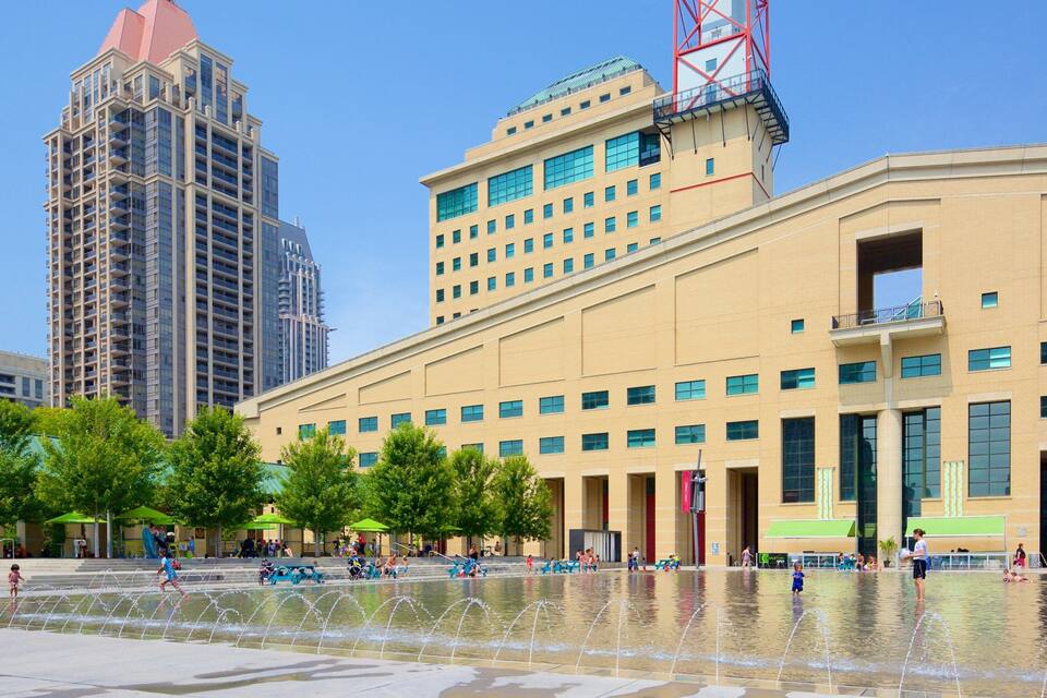 A public square with a water fountain, surrounded by modern buildings and trees.