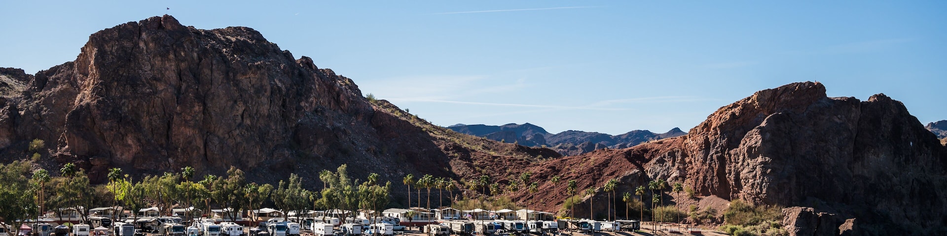 A coastal area with RVs parked along the shore, palm trees, and rocky mountains in the background.