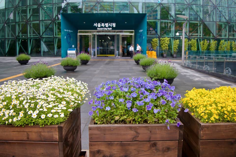A modern building with glass facades, surrounded by flower beds with white, purple, and yellow flowers.