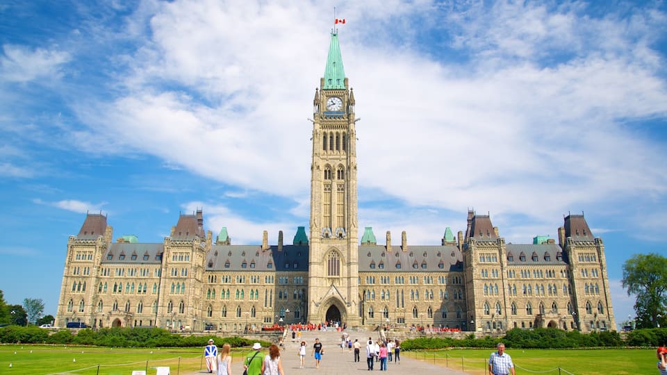 A large historic building with a prominent clock tower, surrounded by a well-maintained lawn and a crowd of people.
