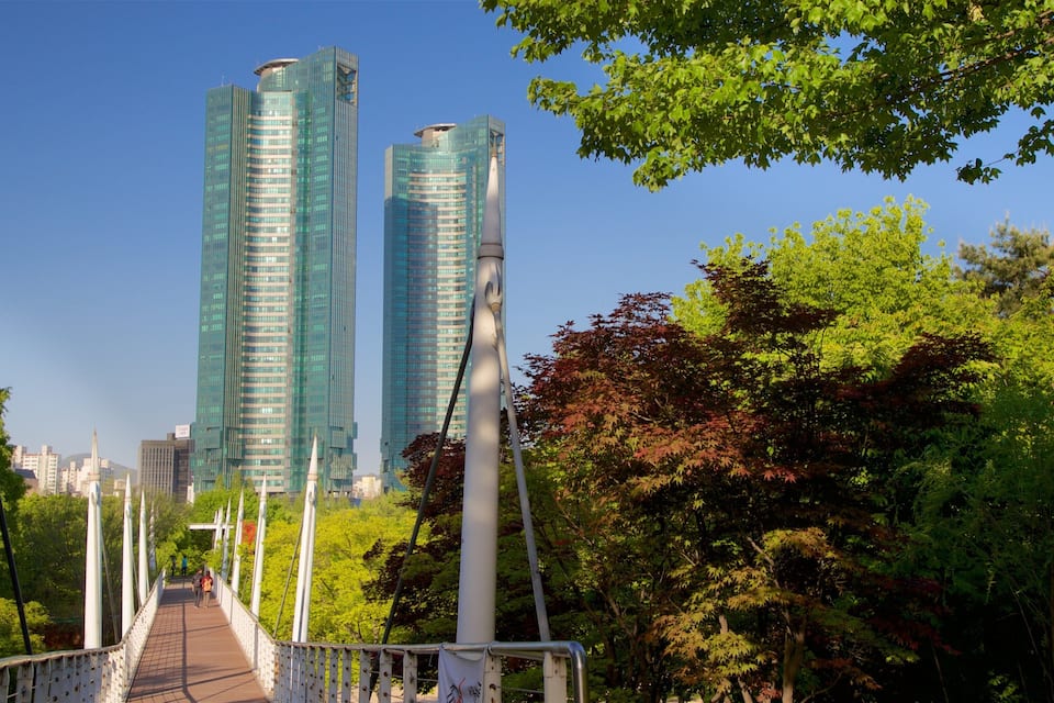 A pedestrian bridge with a view of modern glass buildings and lush green trees.