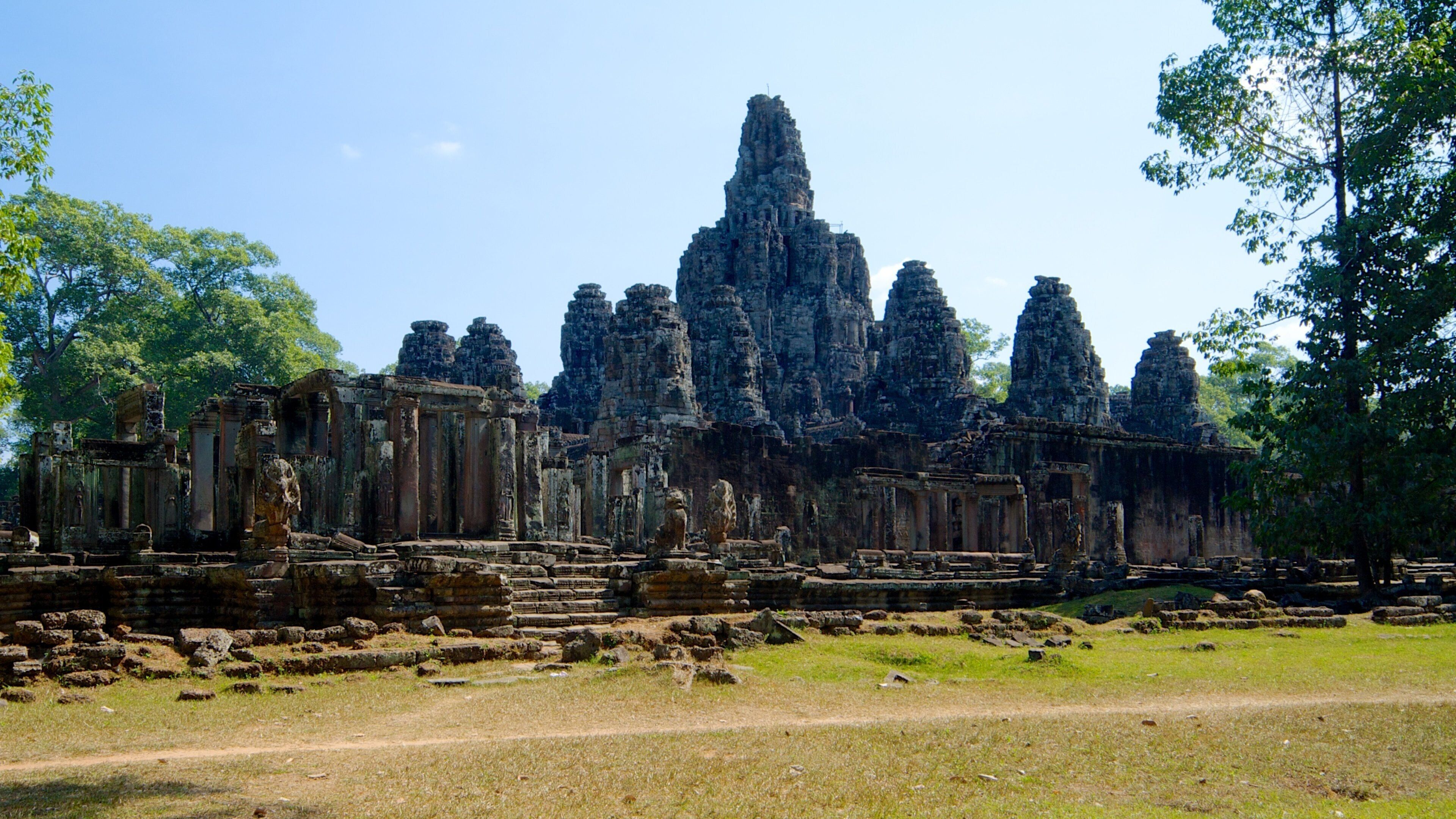 Ancient stone ruins with multiple towers and statues, surrounded by trees and a clear sky.