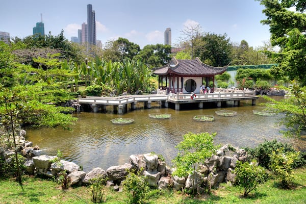Ein traditioneller Pavillon über einem Teich mit Steinen und Grünpflanzen, umgeben von einer Stadtansicht.