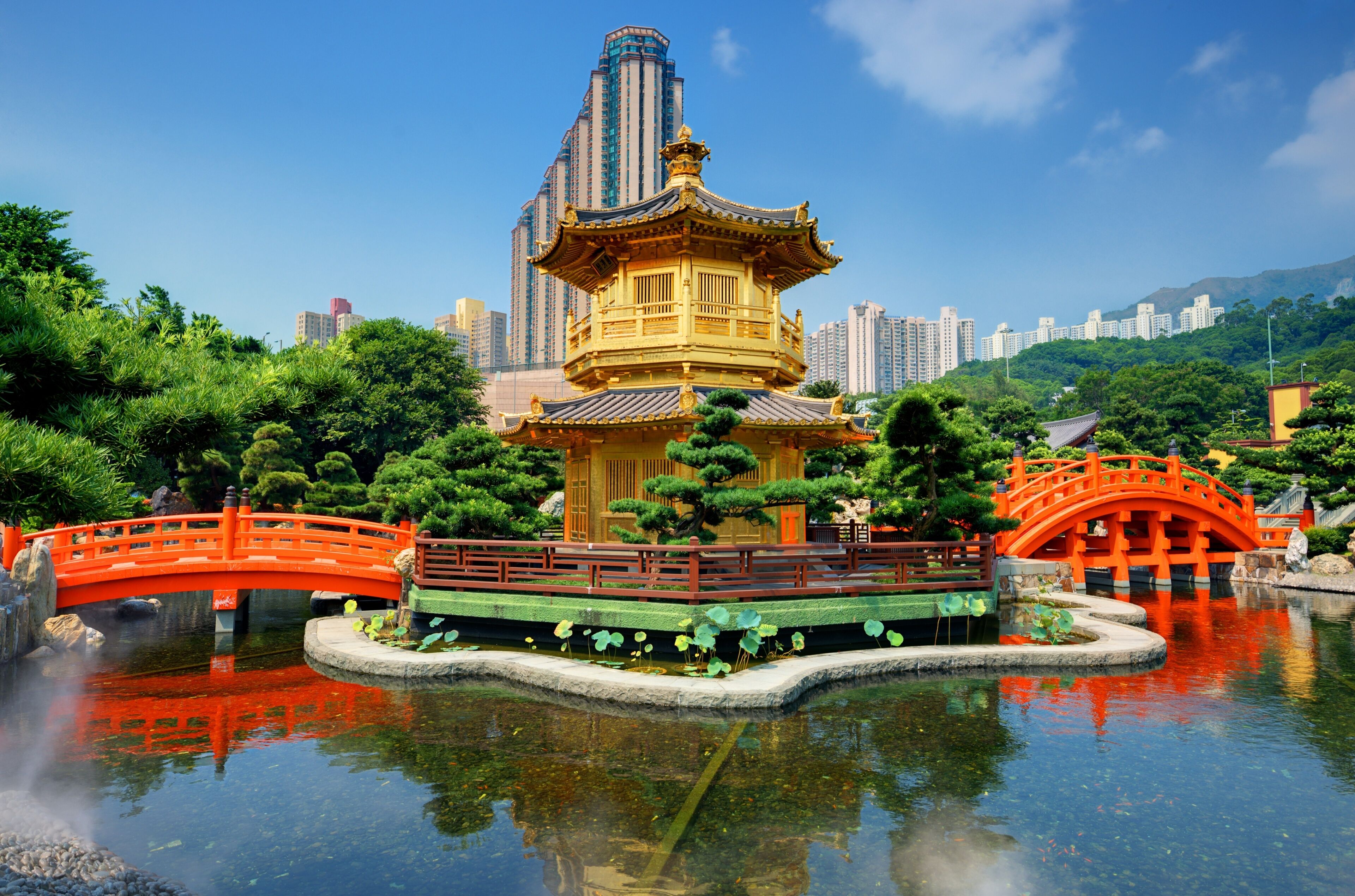 Um pavilhão tradicional chinês com uma ponte vermelha, rodeado por vegetação exuberante e um lago tranquilo.