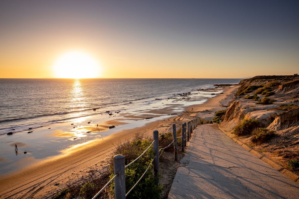 A coastal sunset with a sandy beach, a rocky cliff, and a paved walkway.