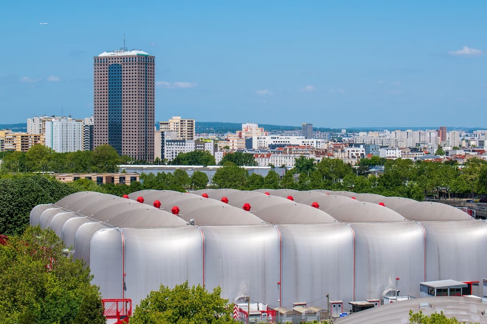 Stadsgezicht met een hoog gebouw, meerdere grote industriële tanks en een heldere blauwe lucht.