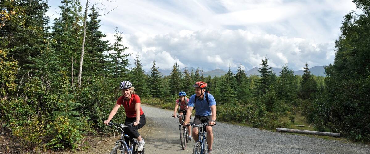 Three cyclists on a gravel road surrounded by dense forest and mountains.