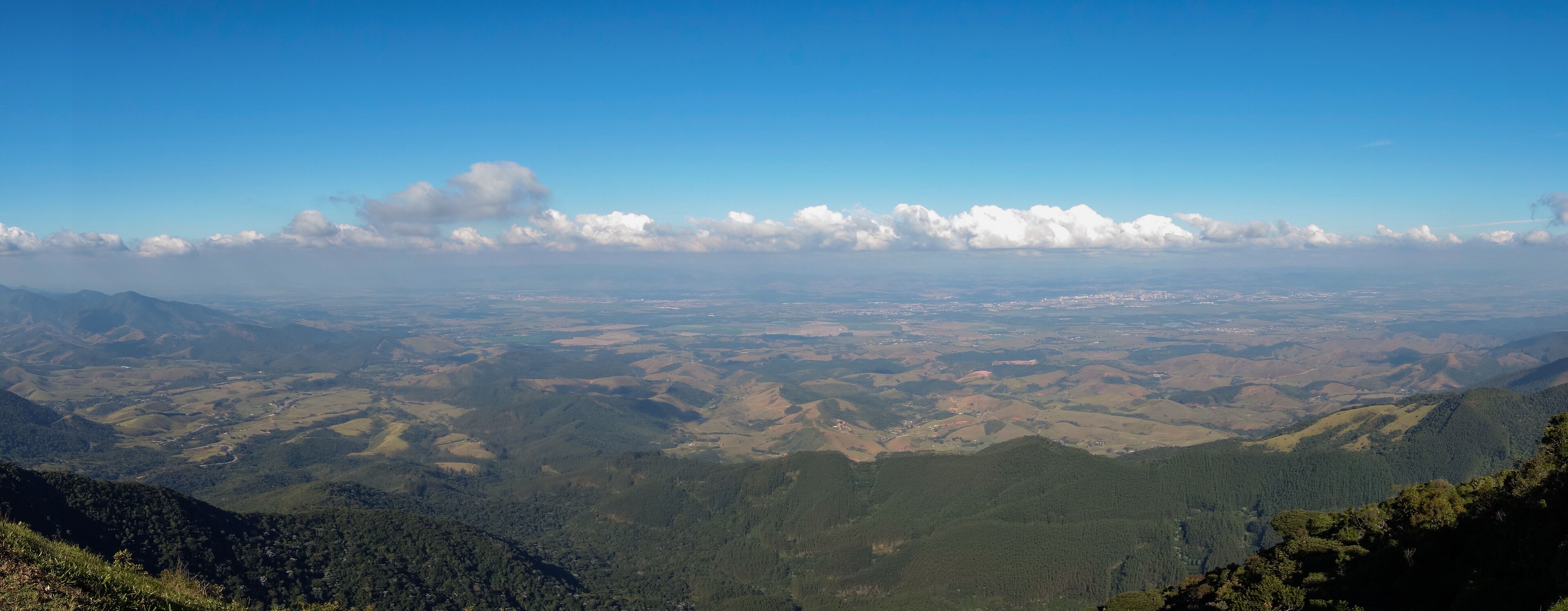 A mountainous landscape with rolling hills, patches of forest, and a clear blue sky with scattered clouds.