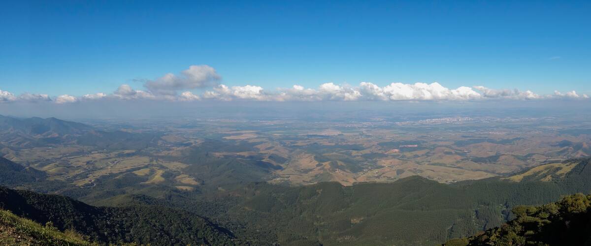 A mountainous landscape with rolling hills, patches of forest, and a clear blue sky with scattered clouds.