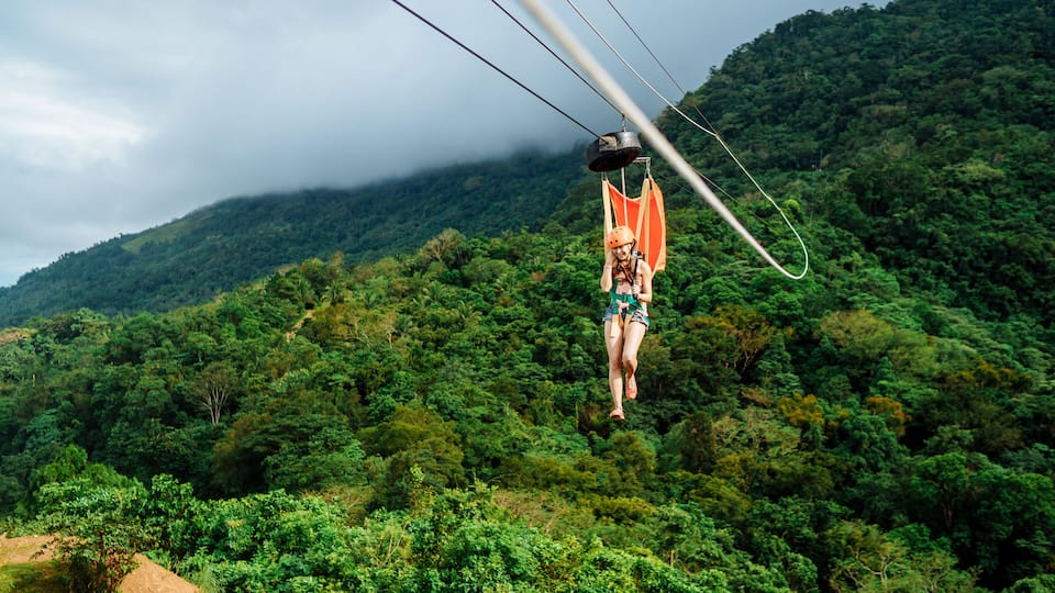 A person suspended in mid-air by a cable, surrounded by lush green forest and mountains.