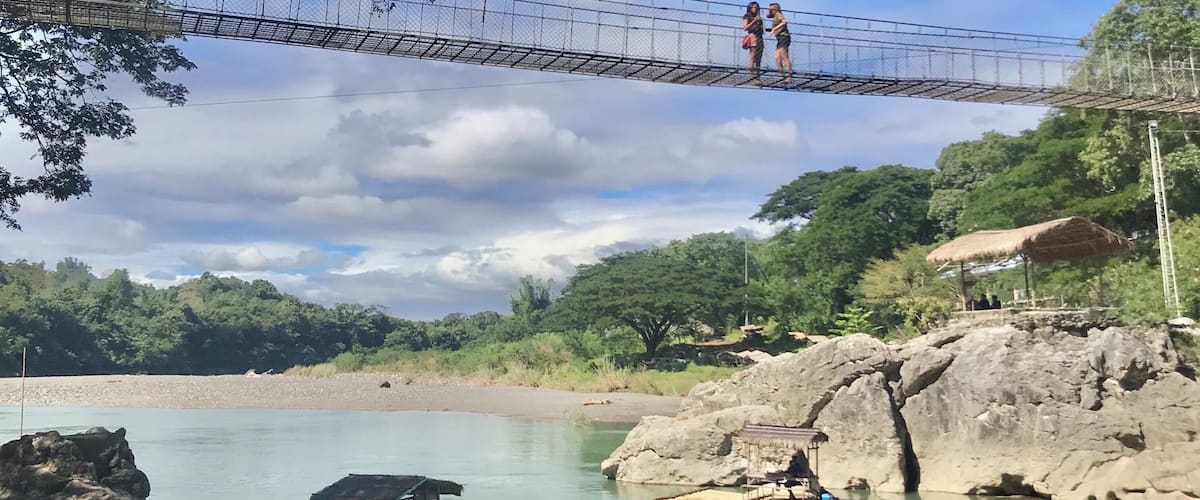 A suspension bridge over a river, surrounded by greenery and rocks.