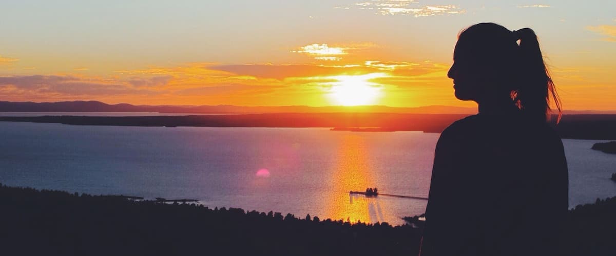 A silhouette of a person overlooking a sunset over a body of water with a boat visible in the distance.