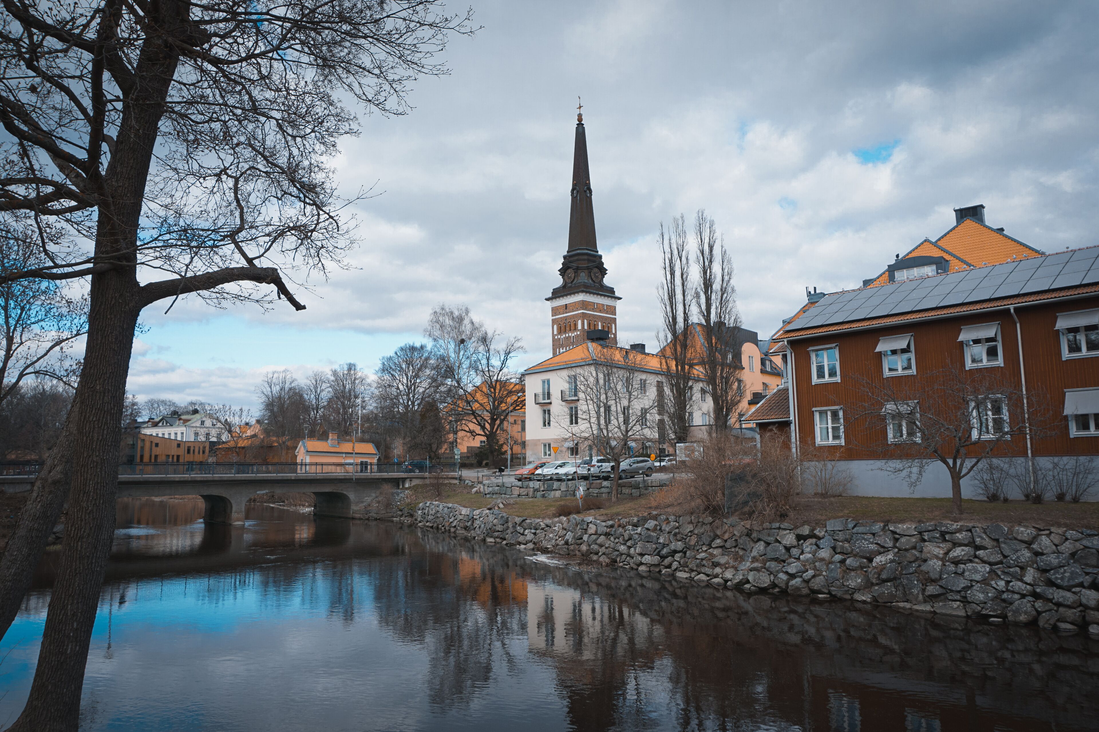 A river with a stone wall, a bridge, a church with a tall spire, and residential buildings.