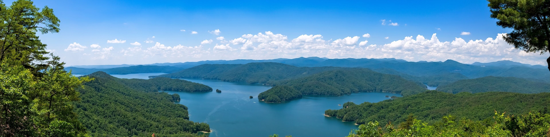 Ein See, umgeben von bewaldeten Hügeln, unter wolkenlosem, blauem Himmel.