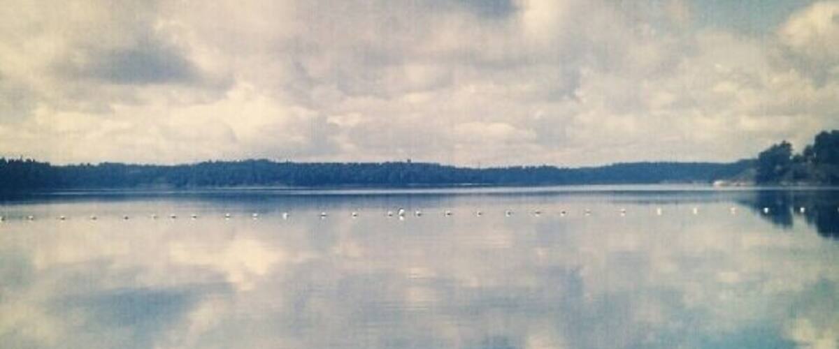 A calm lake with a clear reflection of the sky and clouds, surrounded by a forested shoreline.