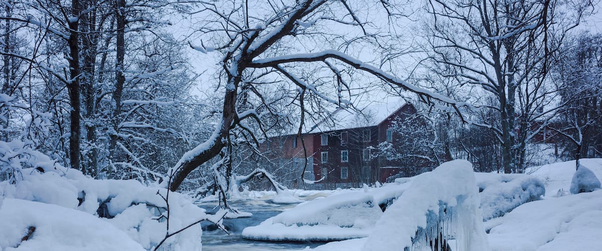 A snow-covered landscape with a frozen stream, bare trees, and a brick building in the background.