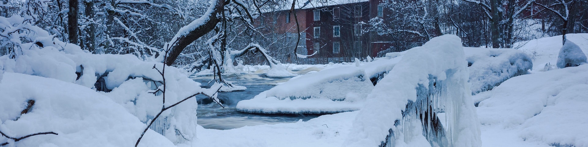A snow-covered landscape with a frozen stream, bare trees, and a brick building in the background.