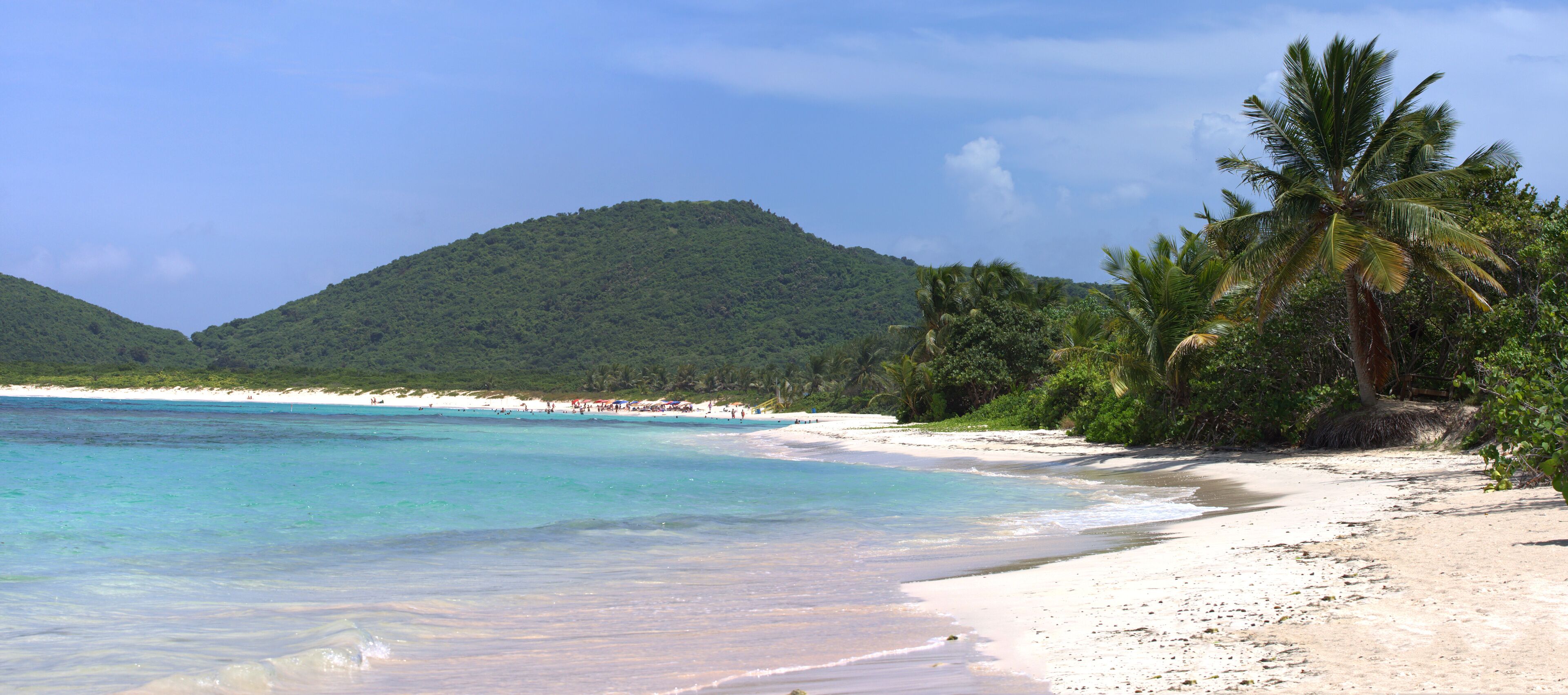 Uma praia com águas turquesa cristalinas, areia branca e colinas verdejantes ao fundo.