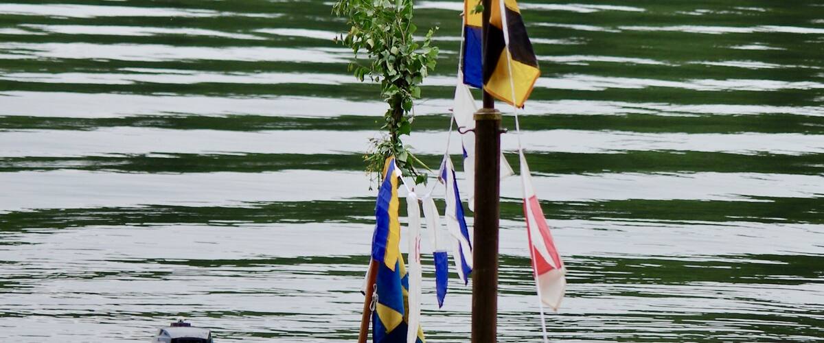 A wooden boat with flags and a small motor, docked by a calm lake with trees in the background.