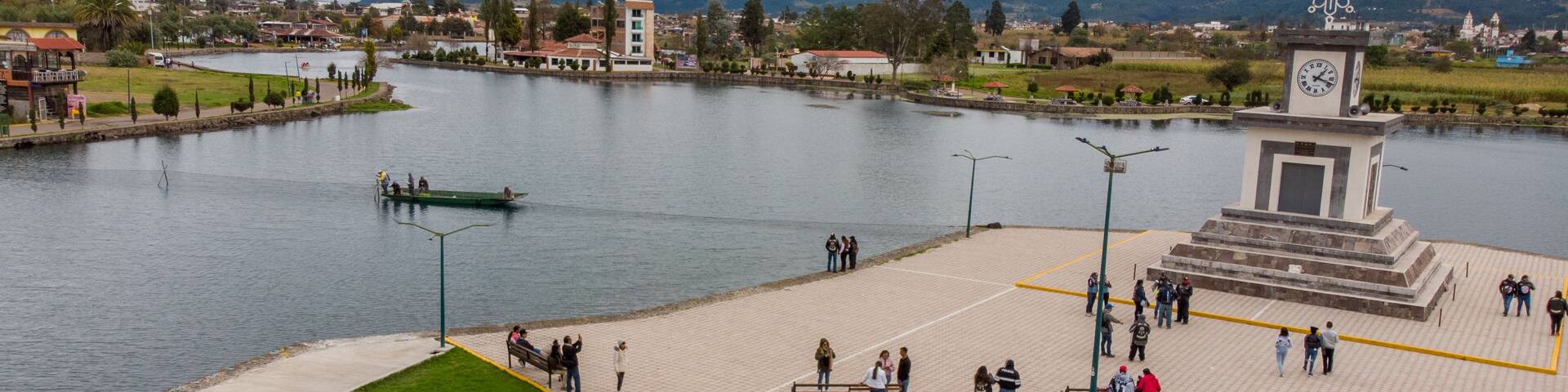 A riverside park with a monument, a boat on the water, and a mountainous backdrop.