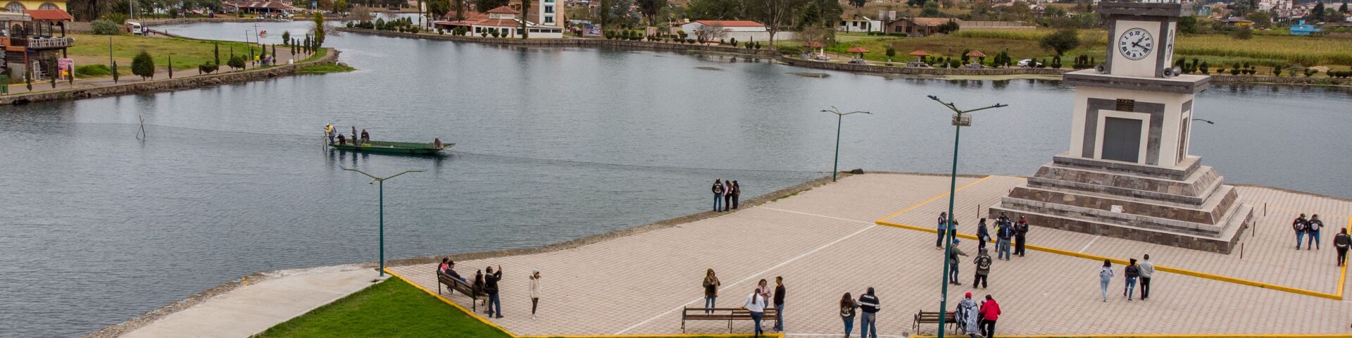 Un parque junto al río con un monumento, un bote en el agua y montañas al fondo.