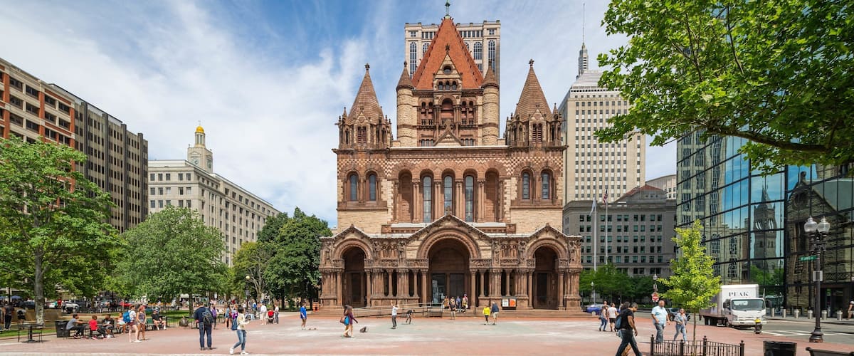 A historic church with a large central tower and multiple spires, surrounded by modern buildings and a public square with people and greenery.