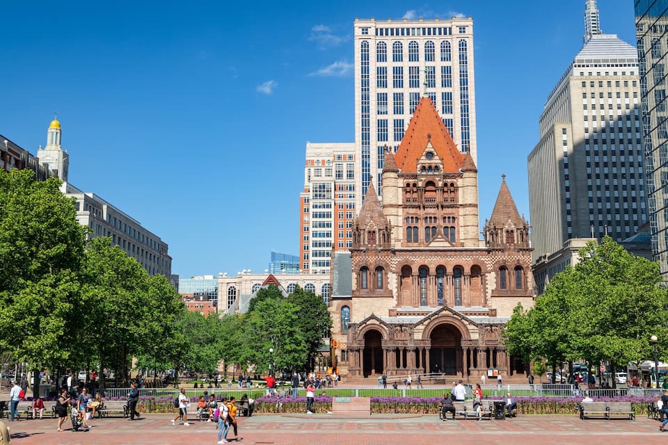 A historic building with a red roof and ornate architecture, surrounded by modern skyscrapers and a public square with people and trees.