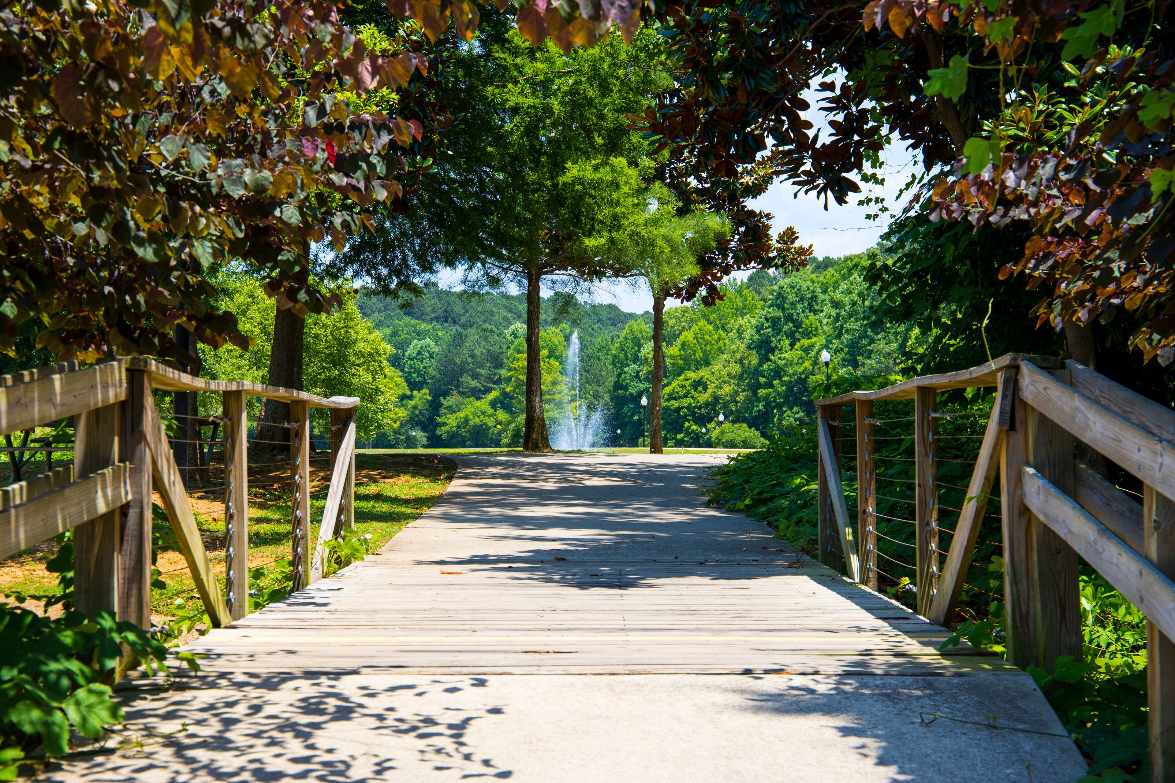 a smooth footpath with a brown wooden hand rail in the park surrounded by a water fountain and lush green trees, grass and plants with blue sky at Logan Farm Park in Acworth Georgia USA