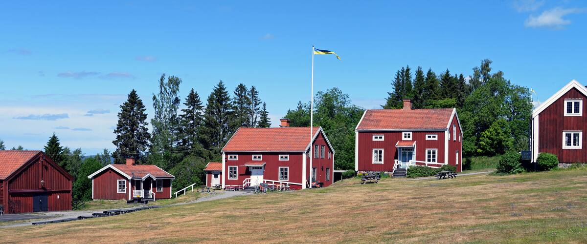 A rural landscape with red wooden houses, a flagpole with a flag, and a clear blue sky.
