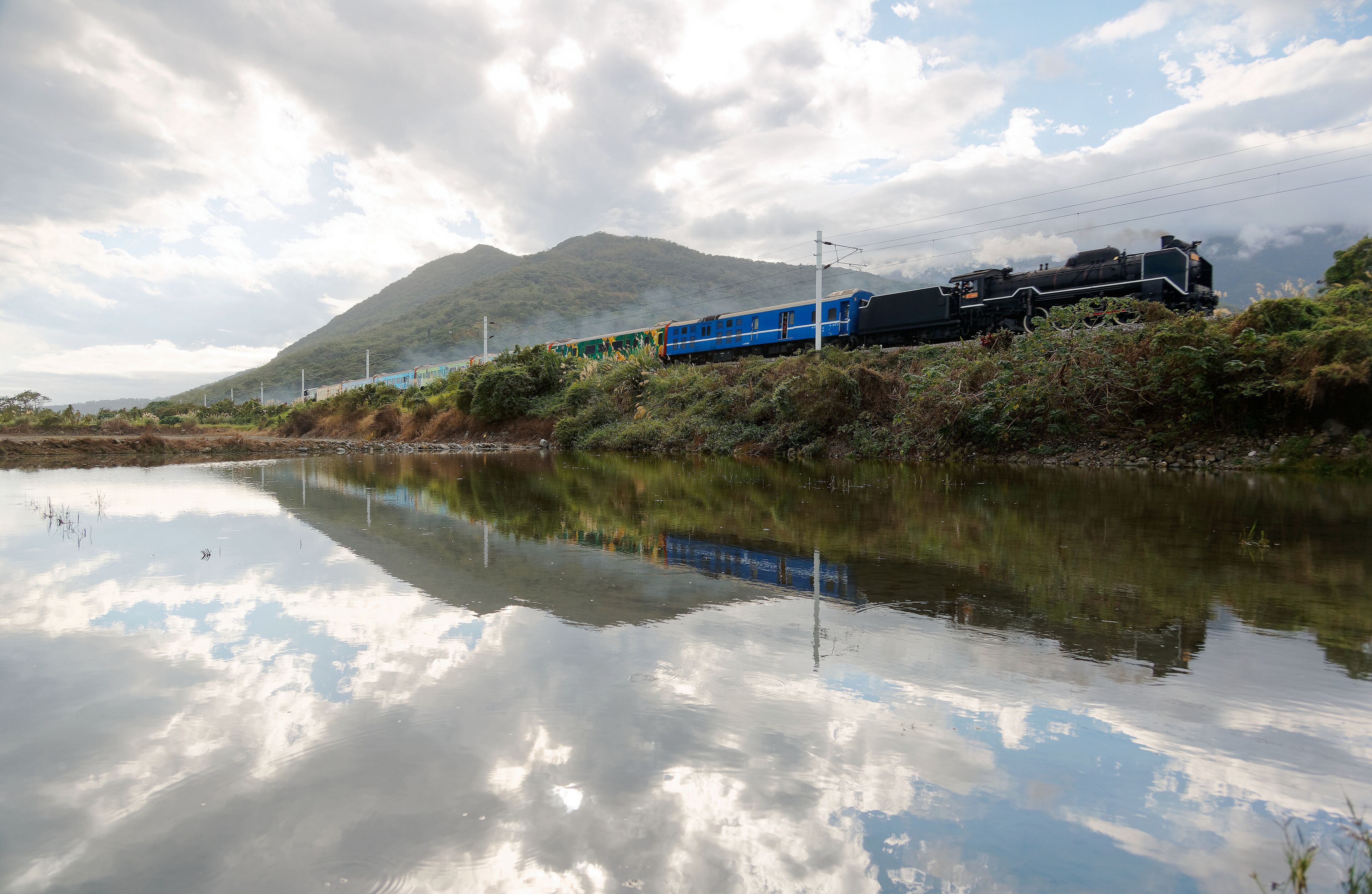 A tourist train hauled by an antique steam locomotive passing by the flooded rice paddies with reflections in the peaceful water on a sunny cloudy summer day in Ji'an Township, Hualien County, Taiwan