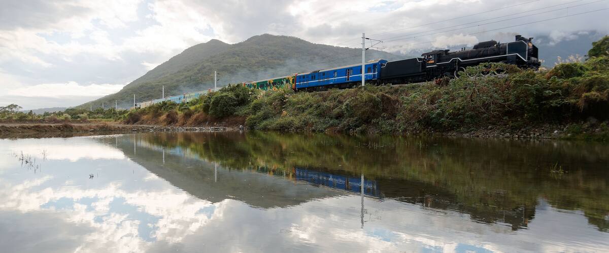 A tourist train hauled by an antique steam locomotive passing by the flooded rice paddies with reflections in the peaceful water on a sunny cloudy summer day in Ji'an Township, Hualien County, Taiwan