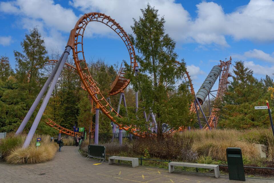 Een achtbaan met een flinke afdaling en een lus, omgeven door bomen en een heldere lucht.