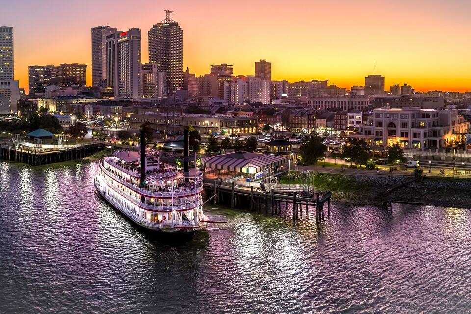 Um barco de passeio atracado em um cais, com um horizonte urbano ao fundo durante o pôr do sol.