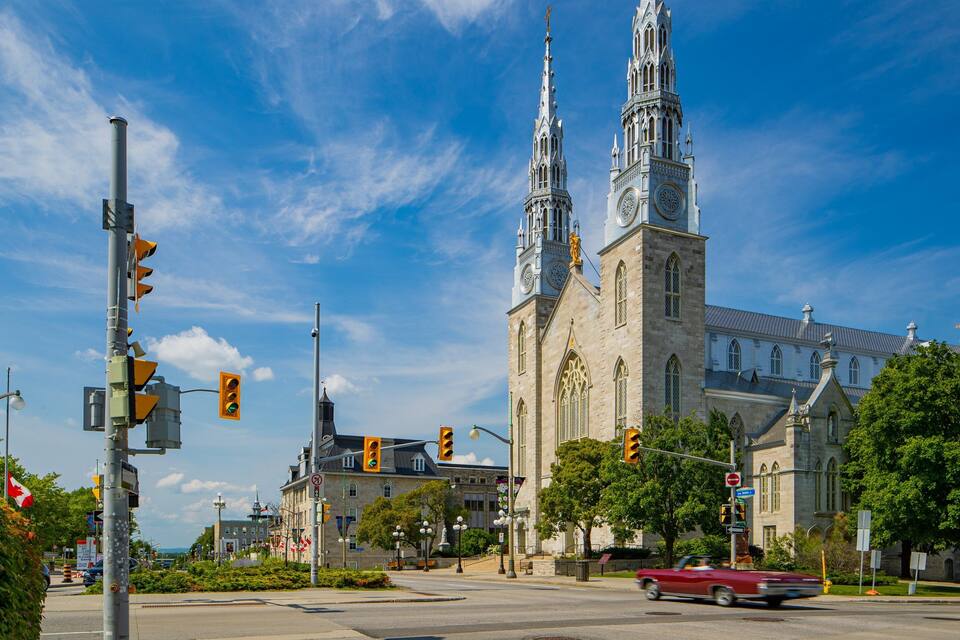 A historic church with twin spires and a clock tower, surrounded by lush greenery and urban street elements.