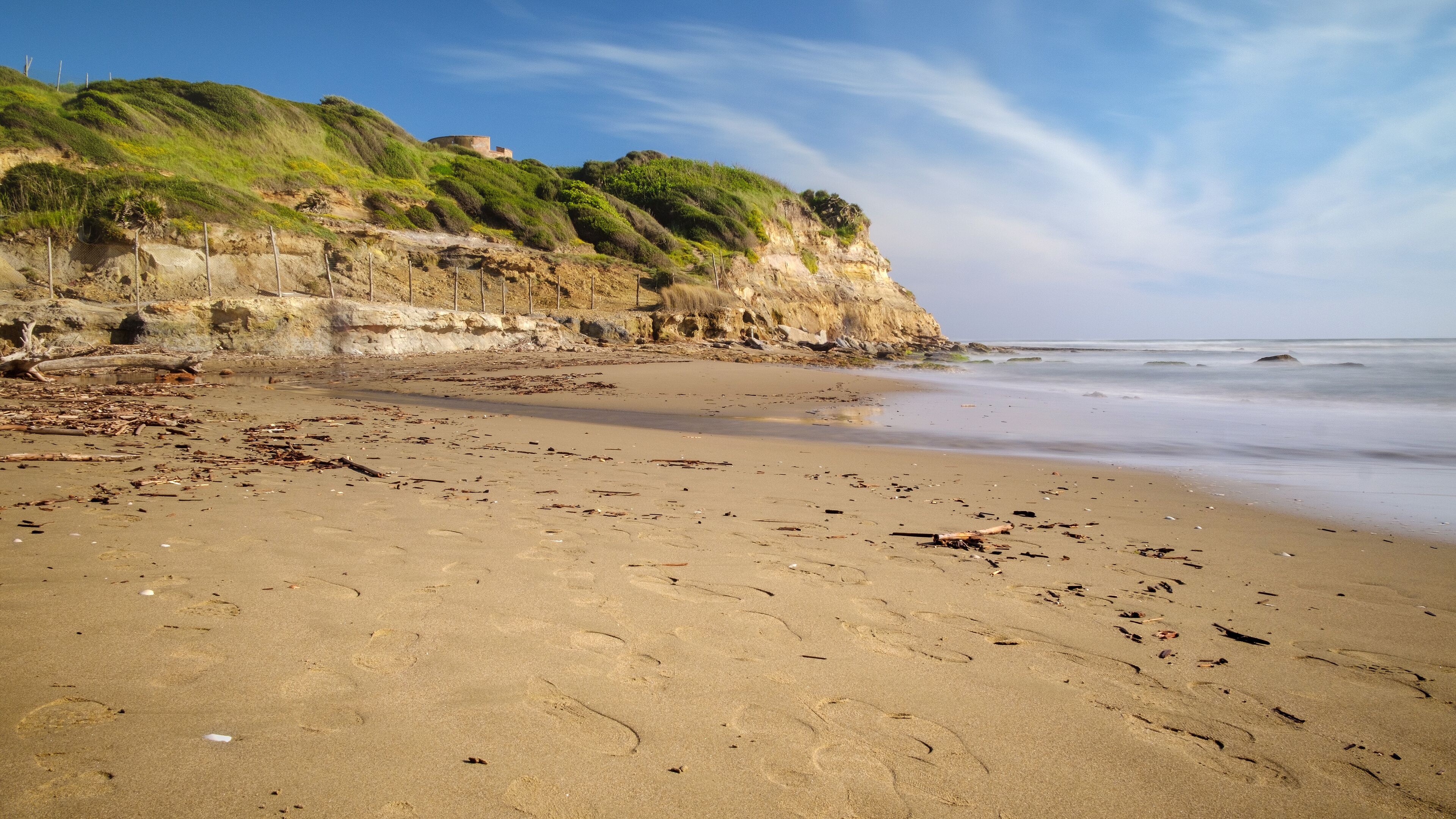A sandy beach with footprints and driftwood, a cliff with greenery, and the ocean in the background.